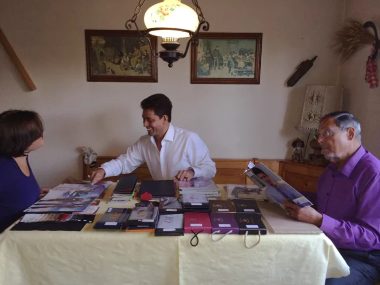 A friendly representative from one of the best tailors in Phuket provides a private consultation for two clients. The table is filled with an extensive selection of fabric swatches and style catalogs, showcasing options for custom bespoke suits and shirts in Phuket.