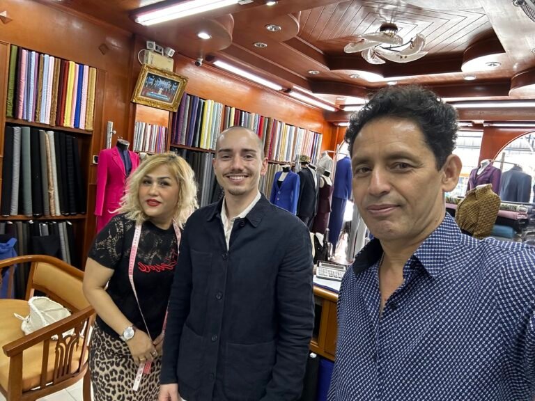 A satisfied customer poses for a photo with the friendly, expert staff at one of the best tailor shops in Phuket. The well-stocked shop, filled with high-quality fabrics for custom suits and bespoke clothing, showcases the premium service offered by this top Phuket tailor.